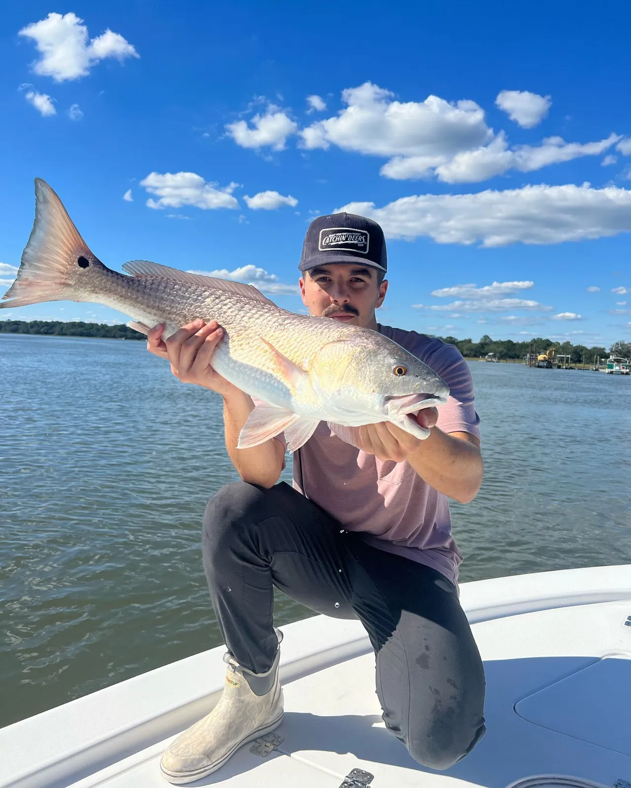 Capt. Dylan Geis kneeling on the bow, holding a big redfish toward the camera under a blue sky